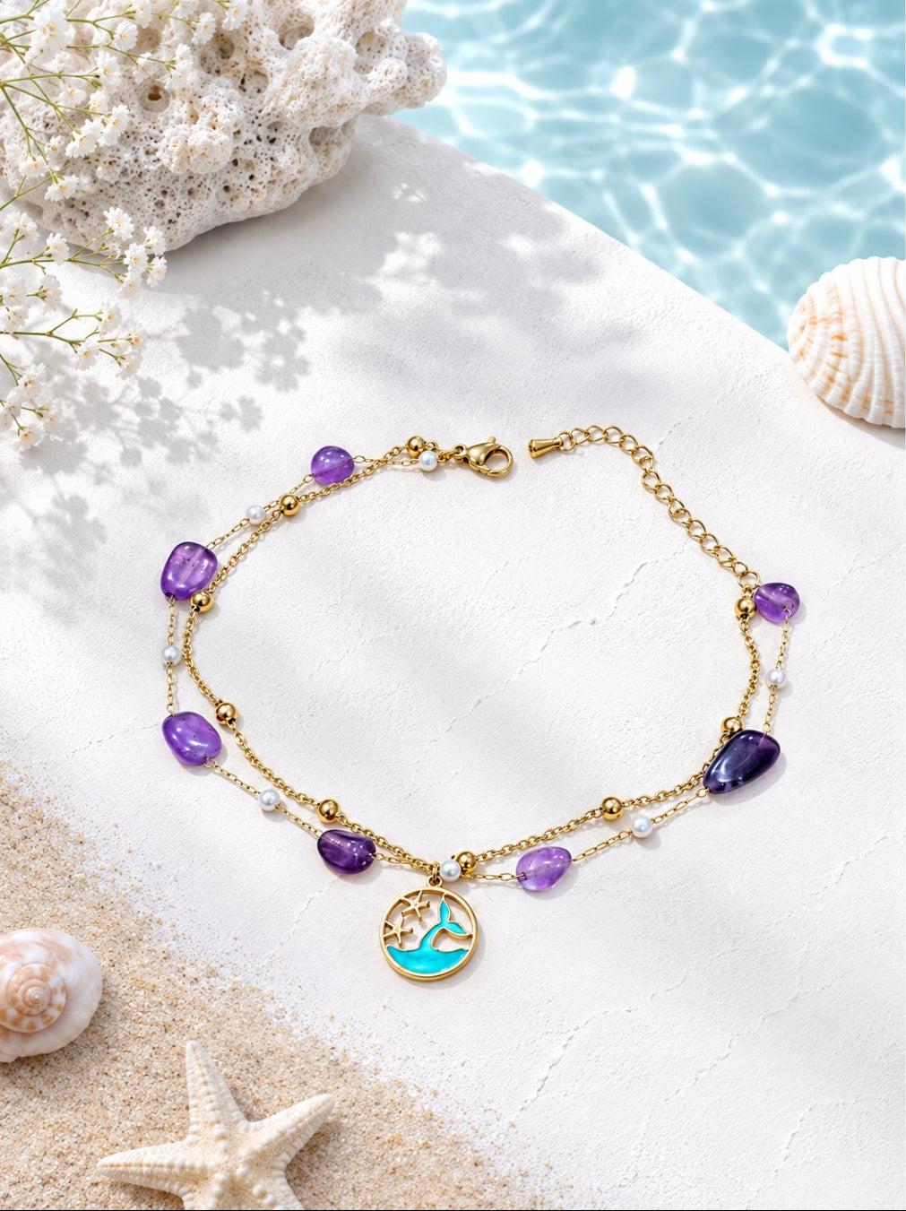 A gold-tone beaded anklet with purple and white beads, a round gold medallion featuring a turquoise and gold wave design, and a small white pearl accent, styled on white sand with seashells and a blue water background.
