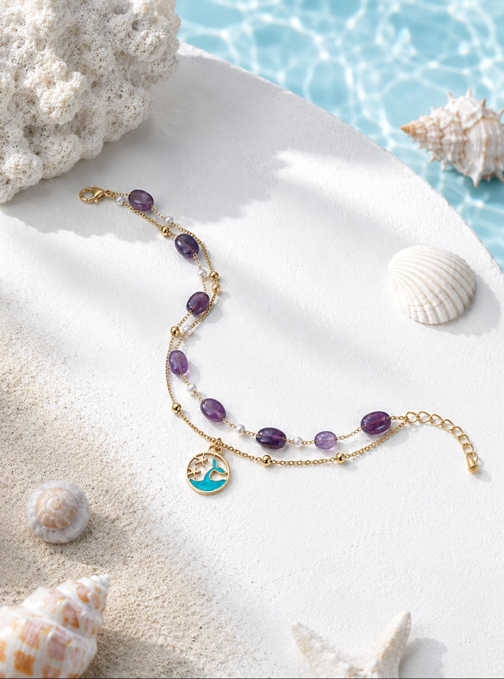A gold-toned beaded bracelet with purple oval beads, clear spacer beads, and a circular turquoise and gold charm, styled on white sand with seashells and a blue water background.