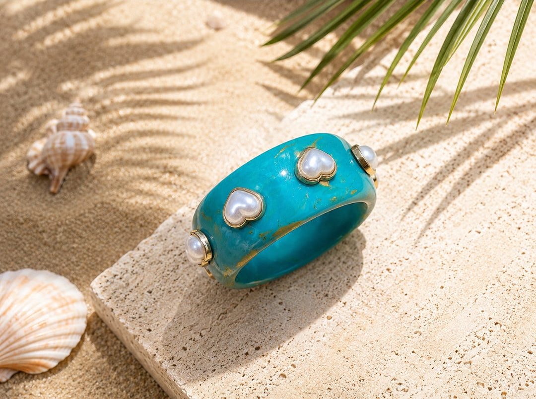 A turquoise bangle bracelet with three heart-shaped pearl inlays and gold-toned trim, displayed on a stone slab over sandy beach props including seashells and palm leaves.