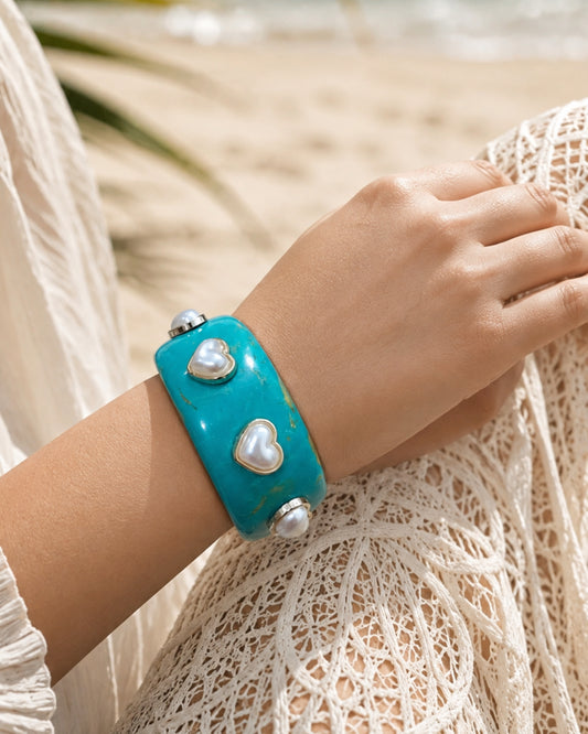 A turquoise cuff bracelet with white faux-pearl heart charms and silver-tone metal accents, worn on a person's wrist in a beach setting.