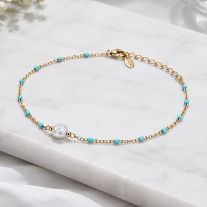 A delicate gold chain bracelet with small turquoise blue beads and a central white pearl, displayed on a white marble slab.