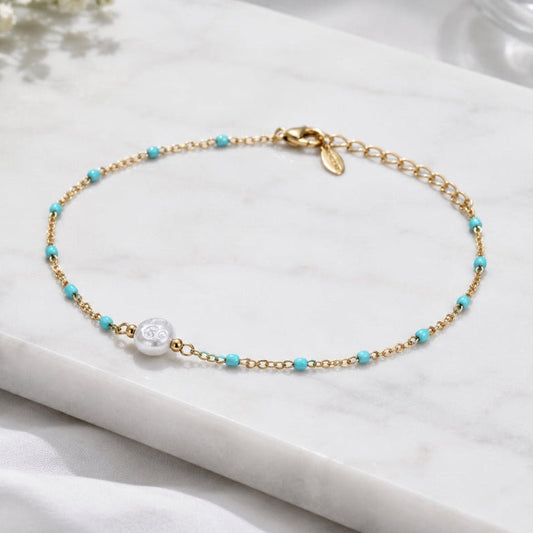A delicate gold chain bracelet with small turquoise blue beads and a central white pearl, displayed on a white marble slab.