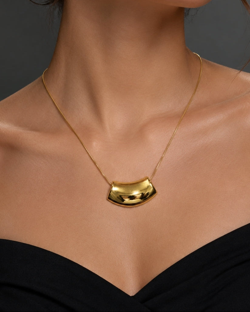 A close-up of a model wearing a gold necklace with a curved, polished pendant against a dark gray background.