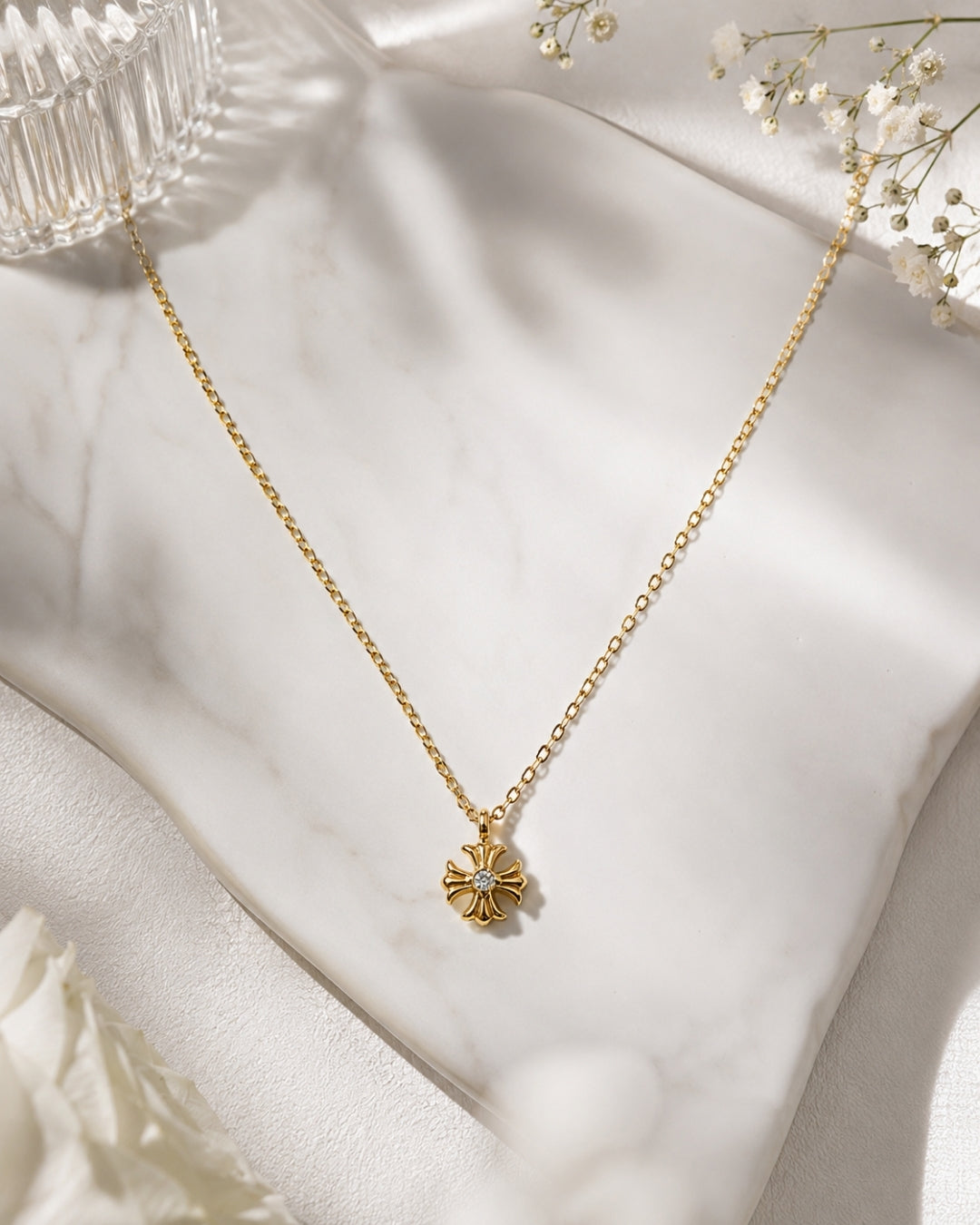 A delicate gold chain necklace with a small four-petal flower pendant, styled on draped white satin fabric with dried baby's breath flowers.