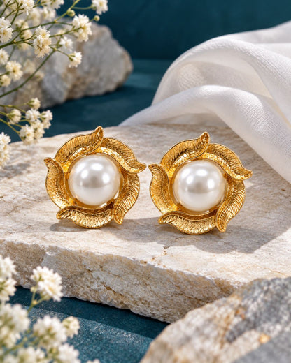 A pair of gold leaf-shaped stud earrings with white pearl centers, displayed on a textured stone with white flowers and draped white fabric.