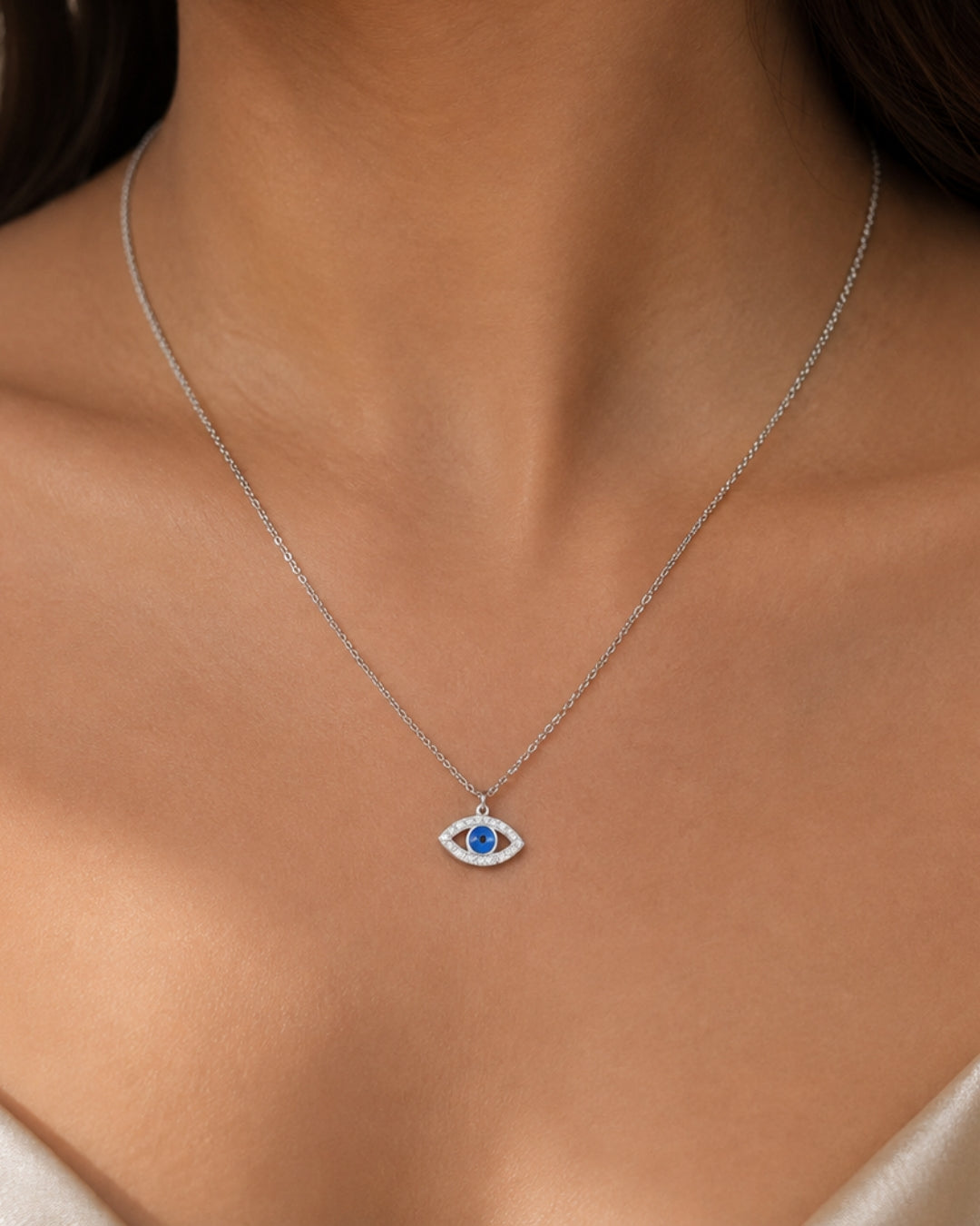 A close-up of a model's neck and chest wearing a delicate silver chain necklace with a small evil eye pendant featuring a blue center.