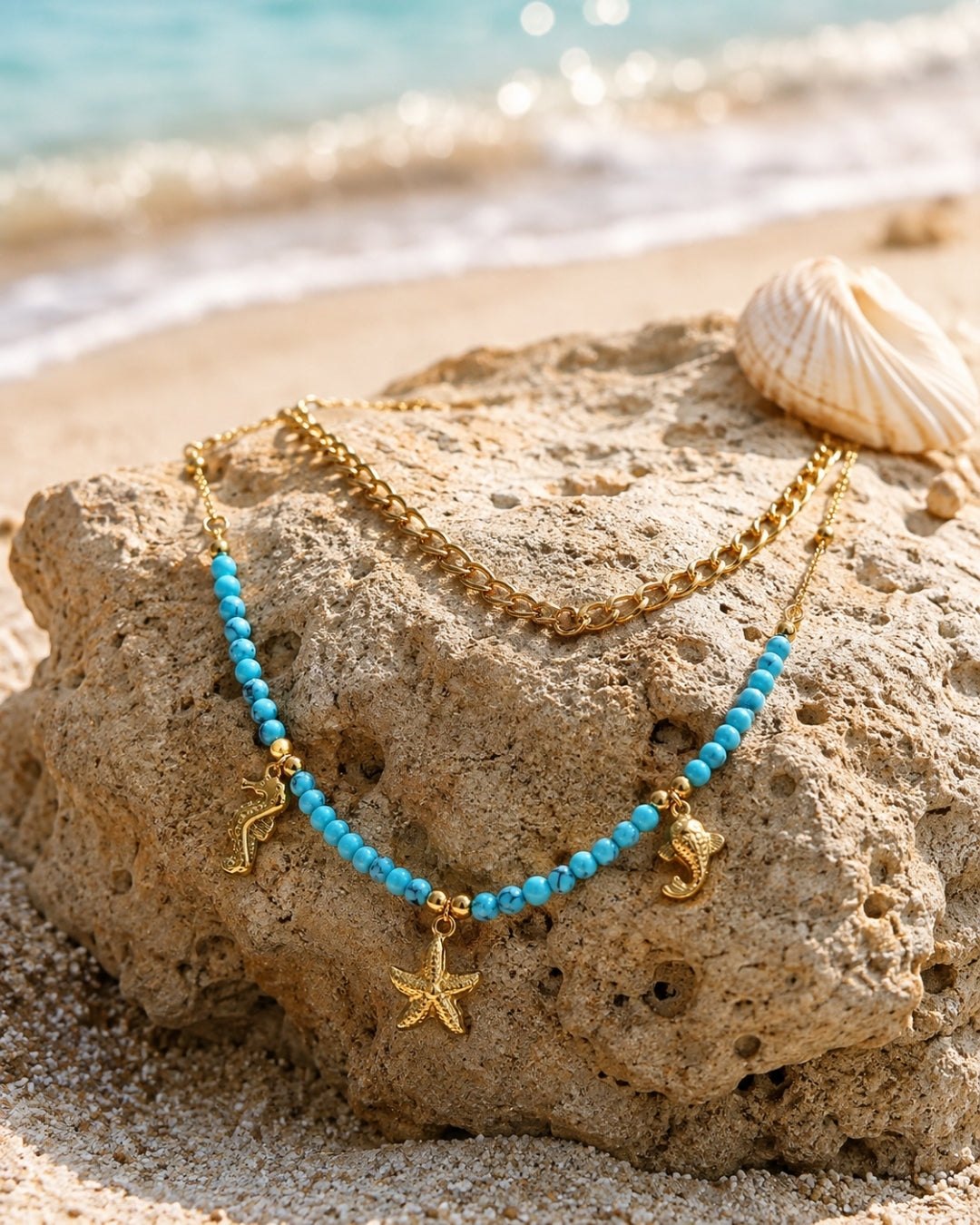 A double-strand necklace displayed on a beach rock, featuring one chain in gold and the other in turquoise beads with gold starfish and seahorse charms, with a seashell and ocean waves in the background.