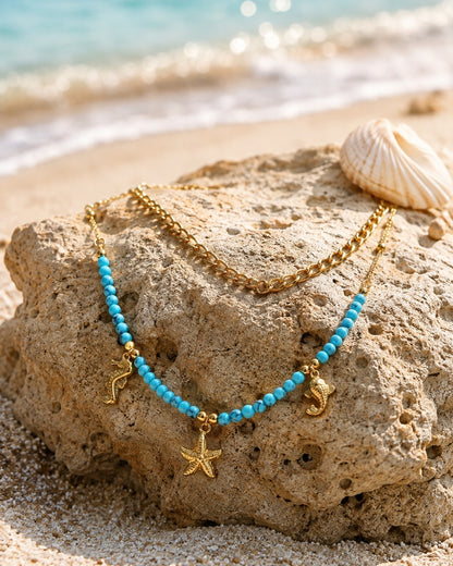 A double-strand necklace displayed on a beach rock, featuring one chain in gold and the other in turquoise beads with gold starfish and seahorse charms, with a seashell and ocean waves in the background.