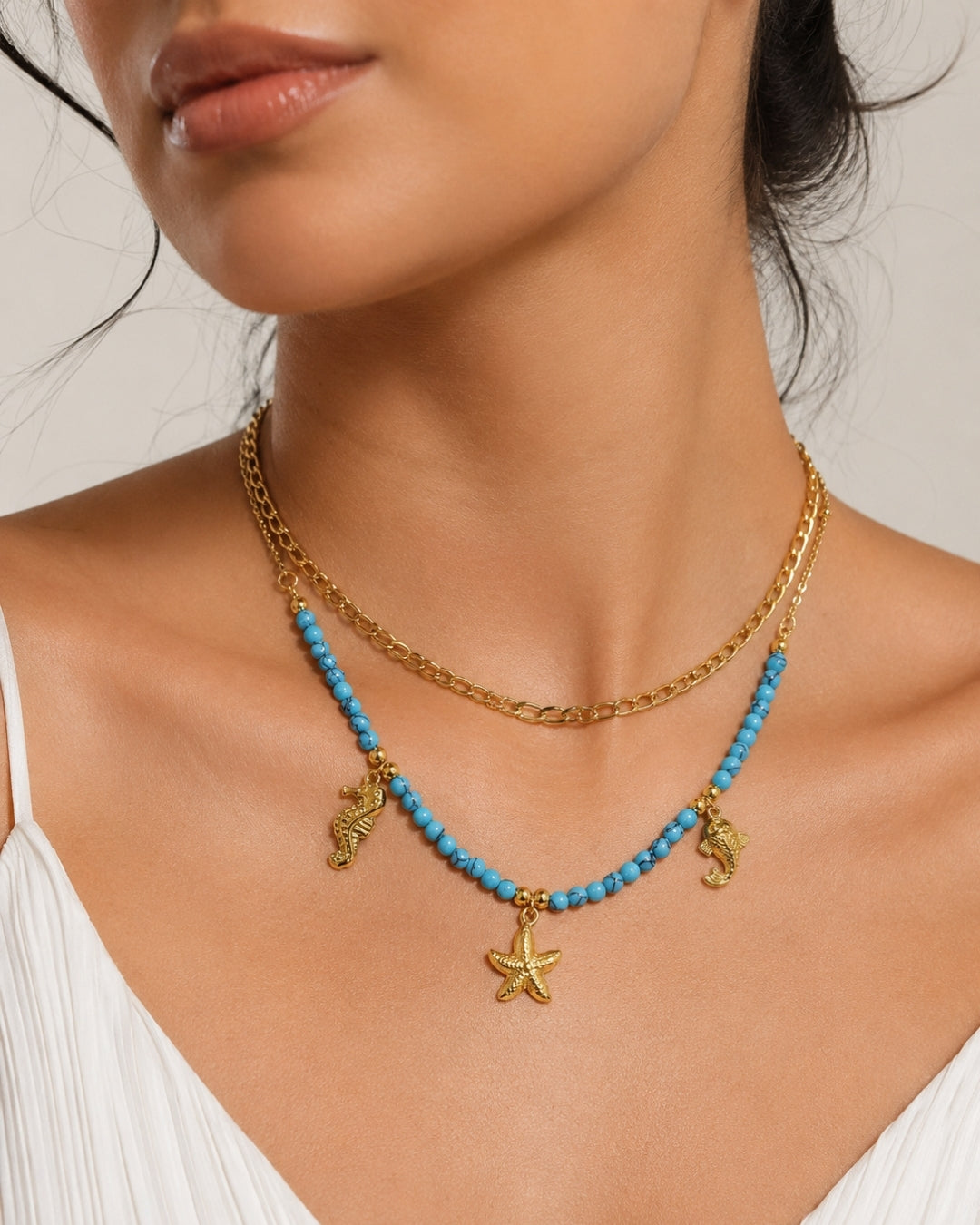 A close-up of a model wearing a double-layered necklace. The top layer is a delicate gold chain, and the bottom layer is a gold chain adorned with small turquoise blue beads and gold seahorse and starfish charms.