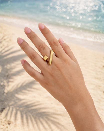 A gold ring with a vertical bar and a small square block detail, worn on a hand resting on a sandy beach with the ocean in the background.
