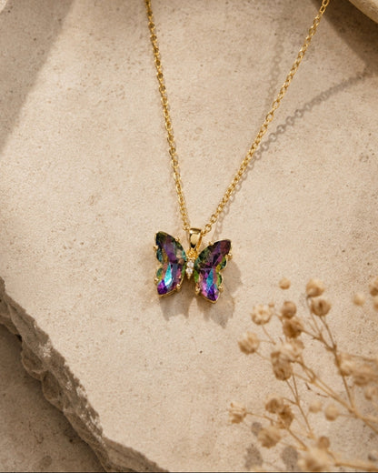 A gold chain necklace with a butterfly pendant featuring iridescent purple, green, and blue gemstones, styled on a textured beige stone surface with dried flowers.