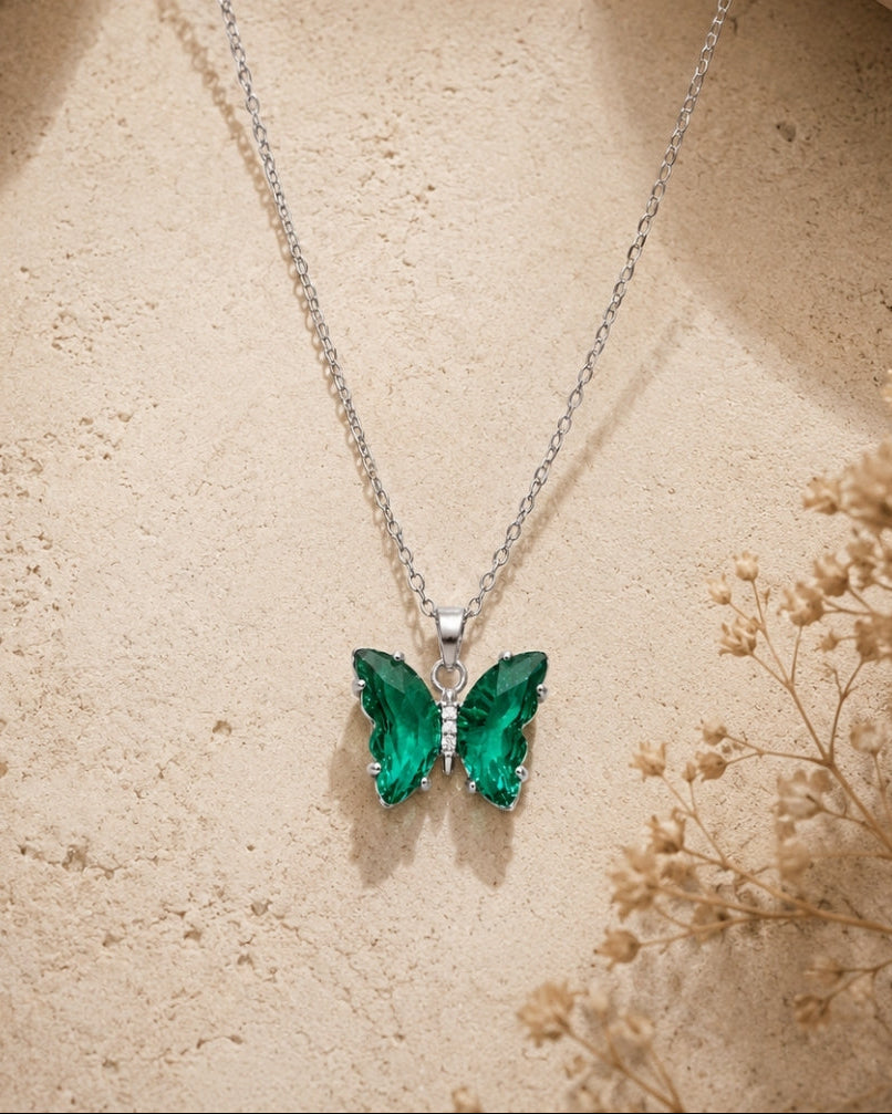 A silver butterfly pendant necklace with vibrant green gemstone wings, displayed on a textured beige stone surface with dried flowers.