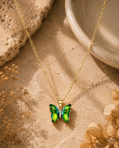 A gold chain necklace with a butterfly pendant featuring vibrant green and blue iridescent wings, styled on a textured beige stone surface with dried flowers and a ceramic dish.