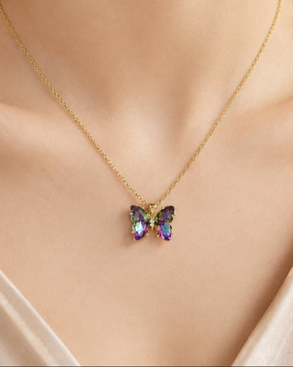A close-up of a model wearing a delicate gold chain necklace with a small butterfly pendant featuring iridescent, multicolored gemstones.