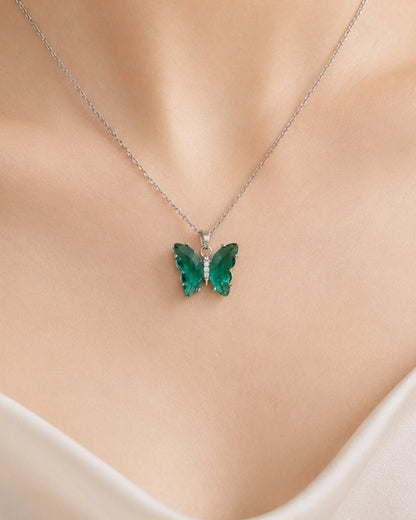 A close-up of a model's neck and chest wearing a delicate silver chain necklace with a small green butterfly pendant.