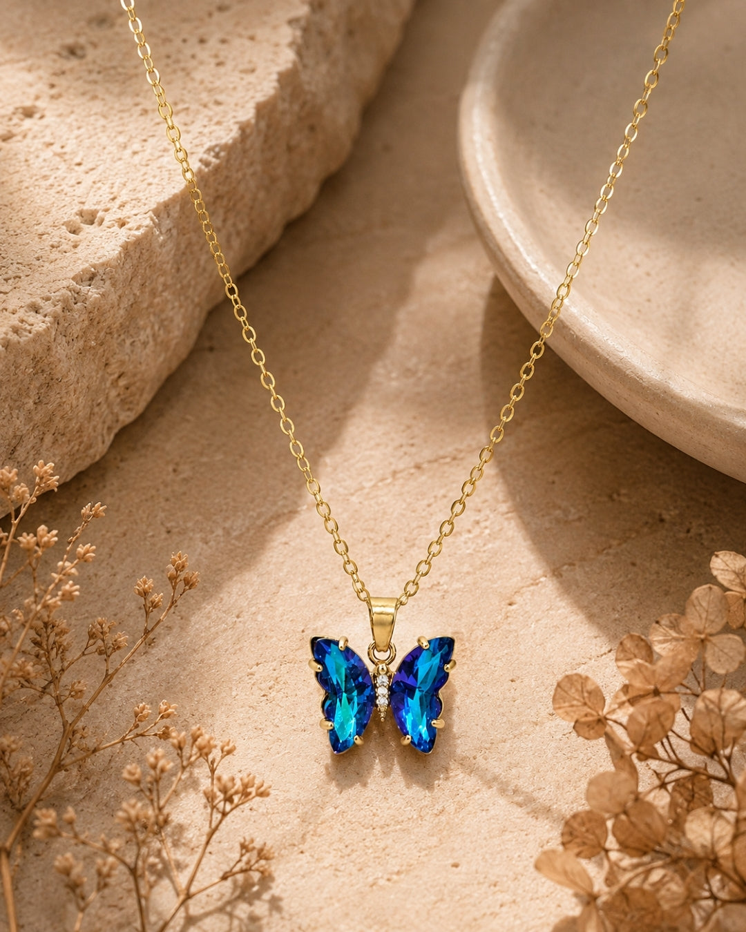 A gold chain necklace with a butterfly-shaped pendant featuring sparkling blue crystals, styled on a textured beige stone surface with dried flowers.
