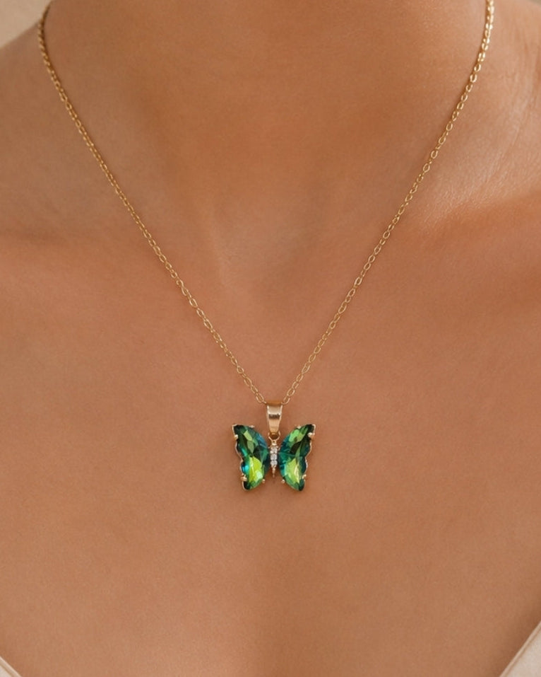 A close-up of a model wearing a delicate gold chain necklace with a butterfly pendant featuring iridescent green and blue crystals, set against a soft beige background.