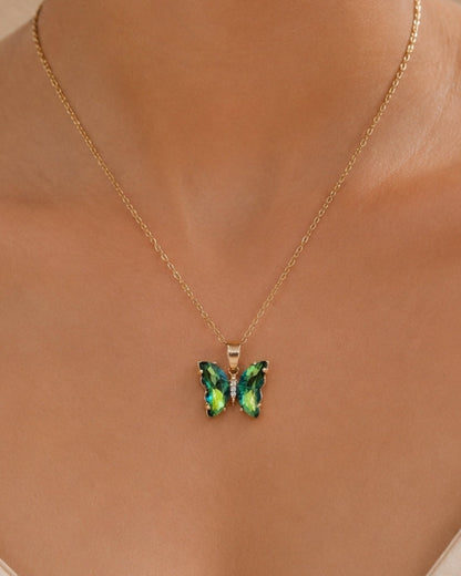 A close-up of a model wearing a delicate gold chain necklace with a butterfly pendant featuring iridescent green and blue crystals, set against a soft beige background.