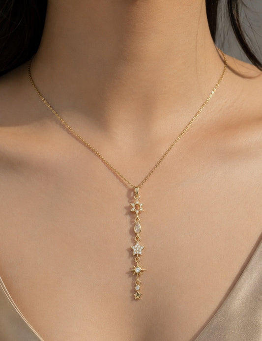 A close-up of a model wearing a delicate gold Y-necklace with a vertical drop of star-shaped charms, some embellished with small clear stones.