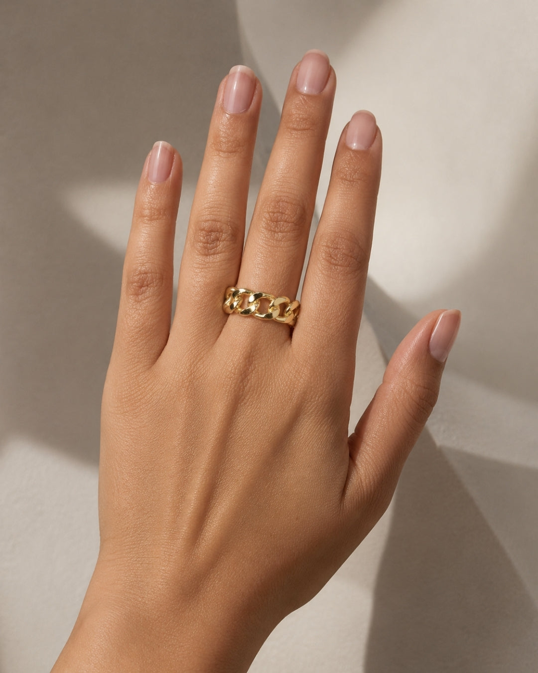 A close-up of a hand wearing a textured gold ring with a raised chain-link design, set against a warm beige background with soft shadows.