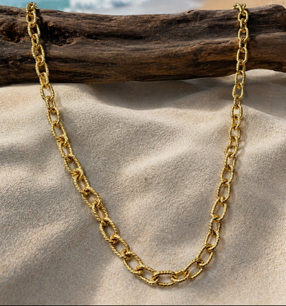 A gold chain necklace draped over a piece of driftwood on a sandy beach, with the ocean and coastline blurred in the background.