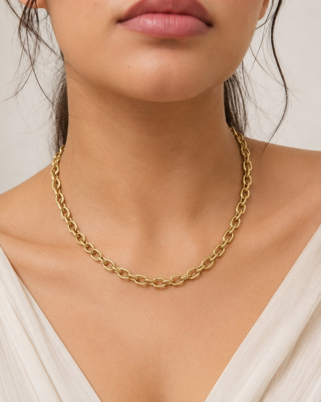 A close-up of a model wearing a delicate gold chain necklace with a textured, slightly wavy link design. The model is wearing a cream-colored top against a soft, light beige background.