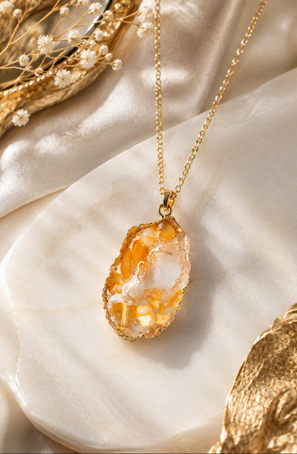 A gold chain necklace with an irregular, raw-cut geode pendant featuring orange and white crystalline formations, styled on draped cream satin fabric with dried flowers.