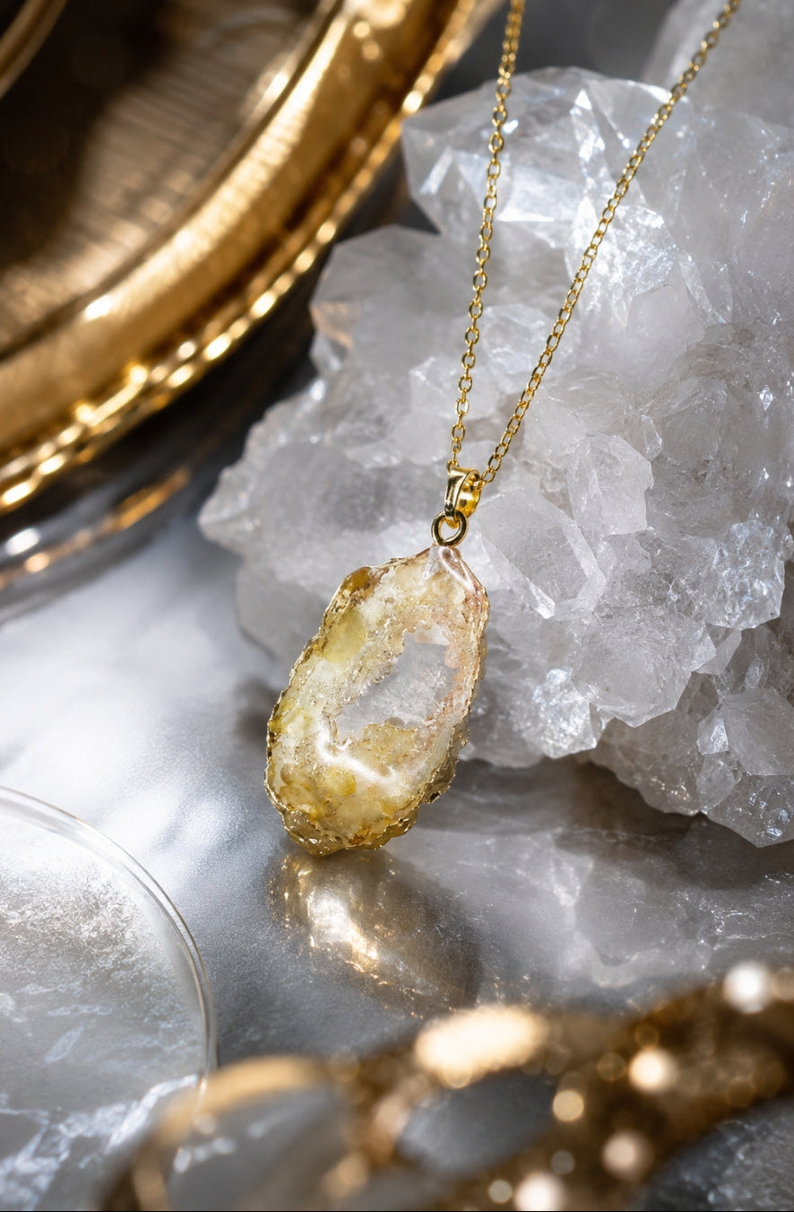 A gold necklace with a raw, irregular-shaped crystal pendant, displayed on a white crystal cluster over a gray marble surface with gold-toned decorative props.