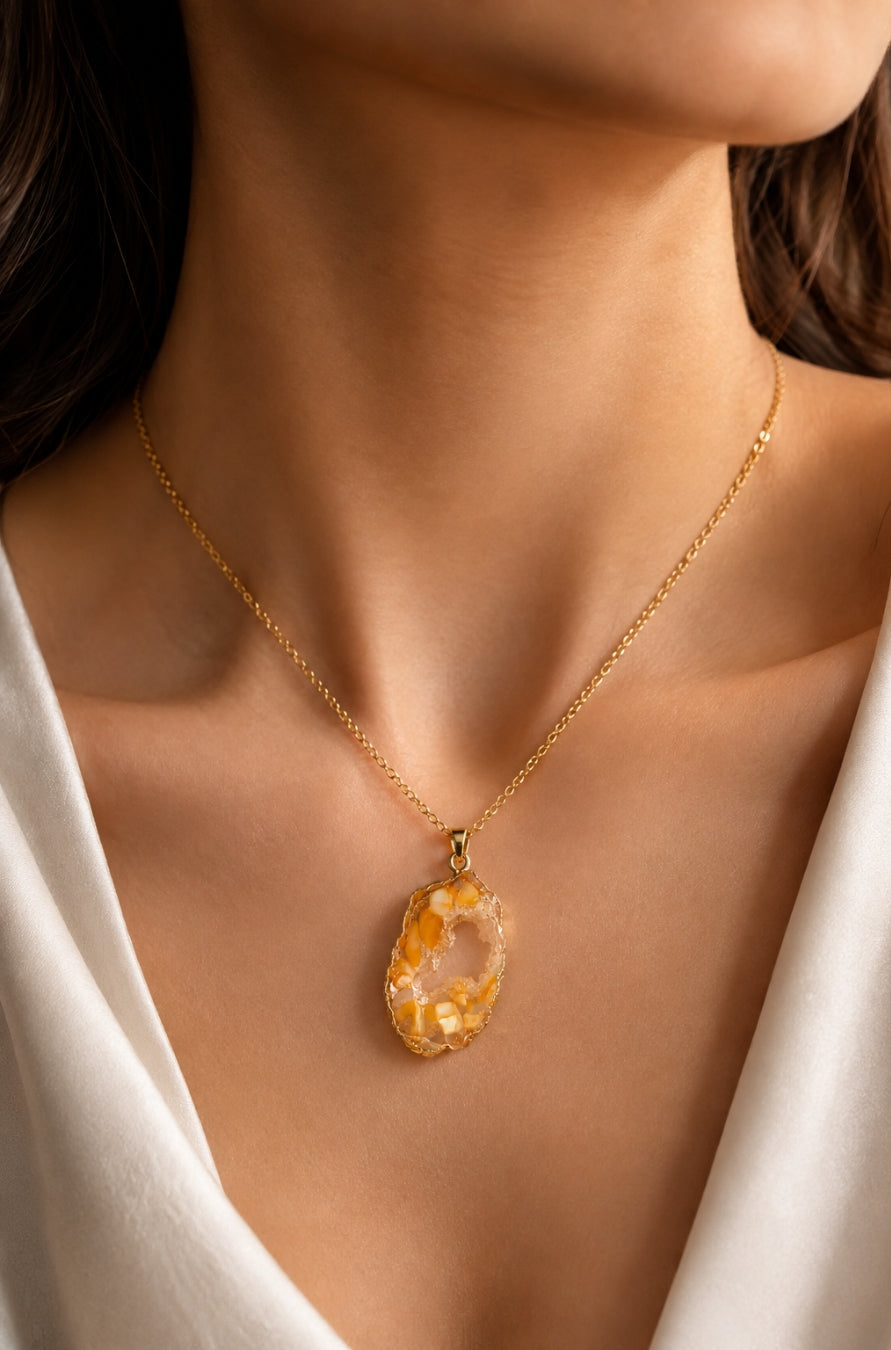 A close-up of a woman's neck and chest wearing a delicate gold chain necklace with a teardrop-shaped, raw, marbled amber-yellow stone pendant.