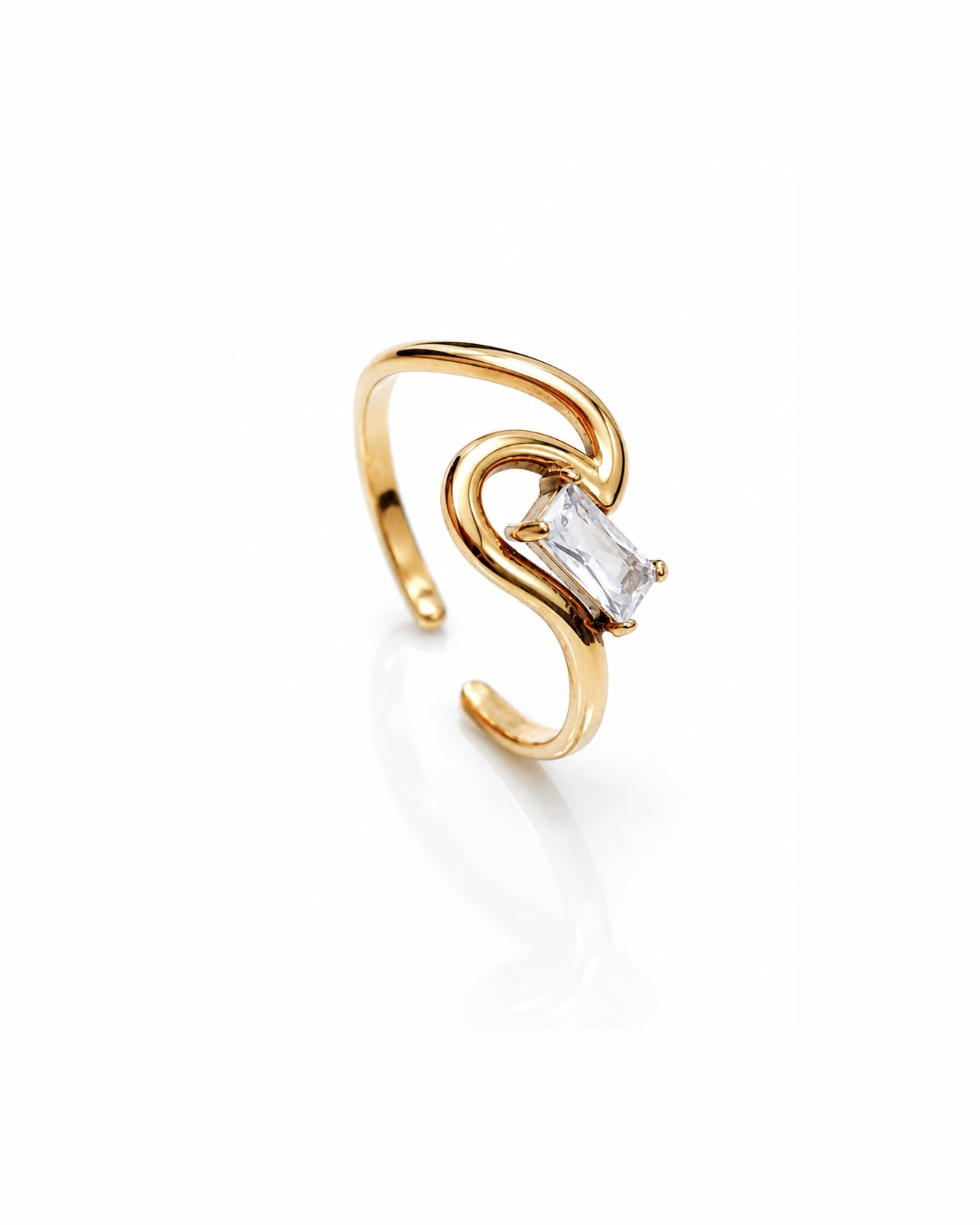 A gold-plated open ring with a curved, sculptural design and a clear rectangular baguette gemstone, shown on a reflective white surface.