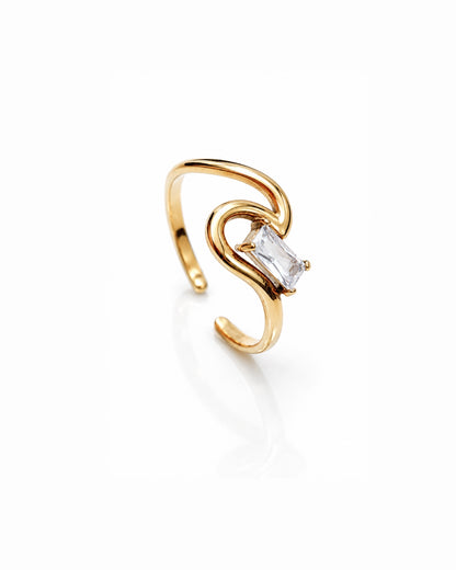 A gold-plated open ring with a curved, sculptural design and a clear rectangular baguette gemstone, shown on a reflective white surface.