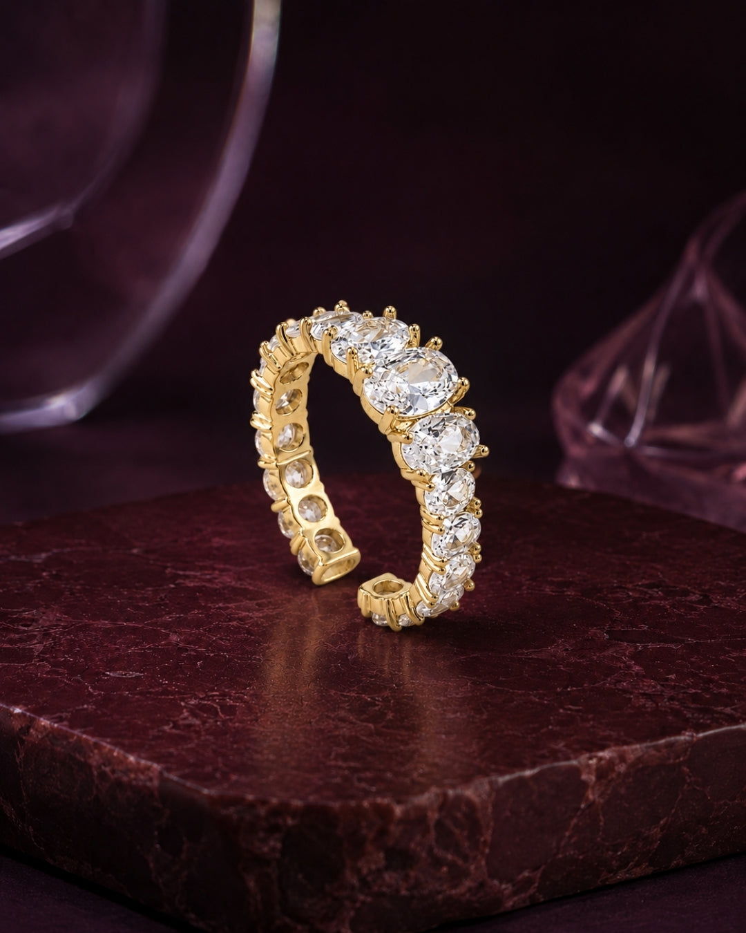 A yellow gold eternity ring set with round brilliant-cut diamonds, displayed on a dark brown marble block against a dark purple background.