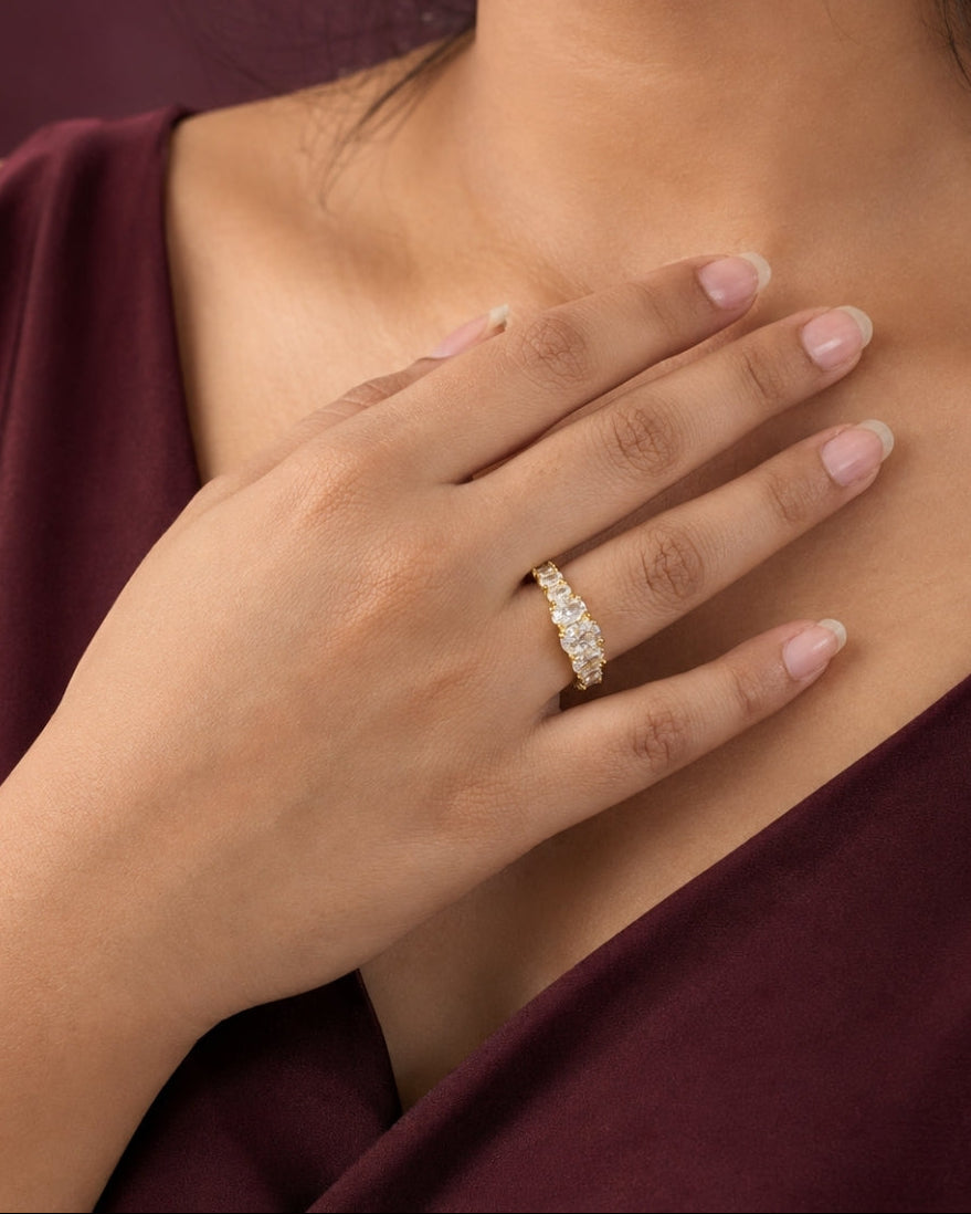 A close-up lifestyle shot of a woman wearing a delicate gold band ring adorned with small clear stones, resting her hand on her chest. She is wearing a deep burgundy top against a matching dark background.