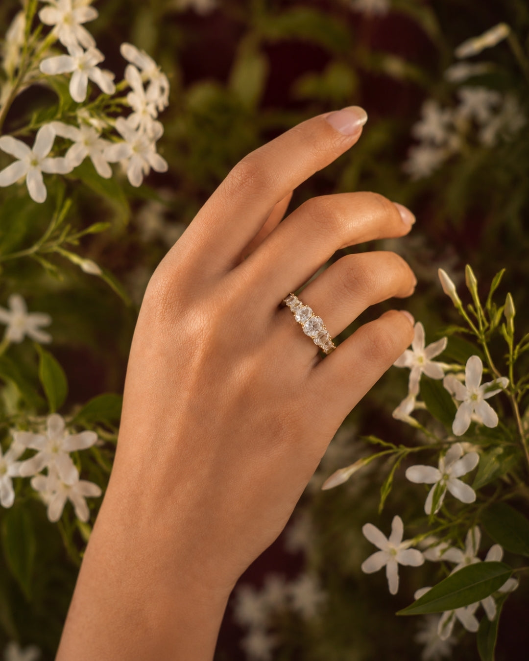 A close-up of a hand wearing a delicate silver ring with a row of clear stones, set against a soft-focus background of green leaves and small white flowers.