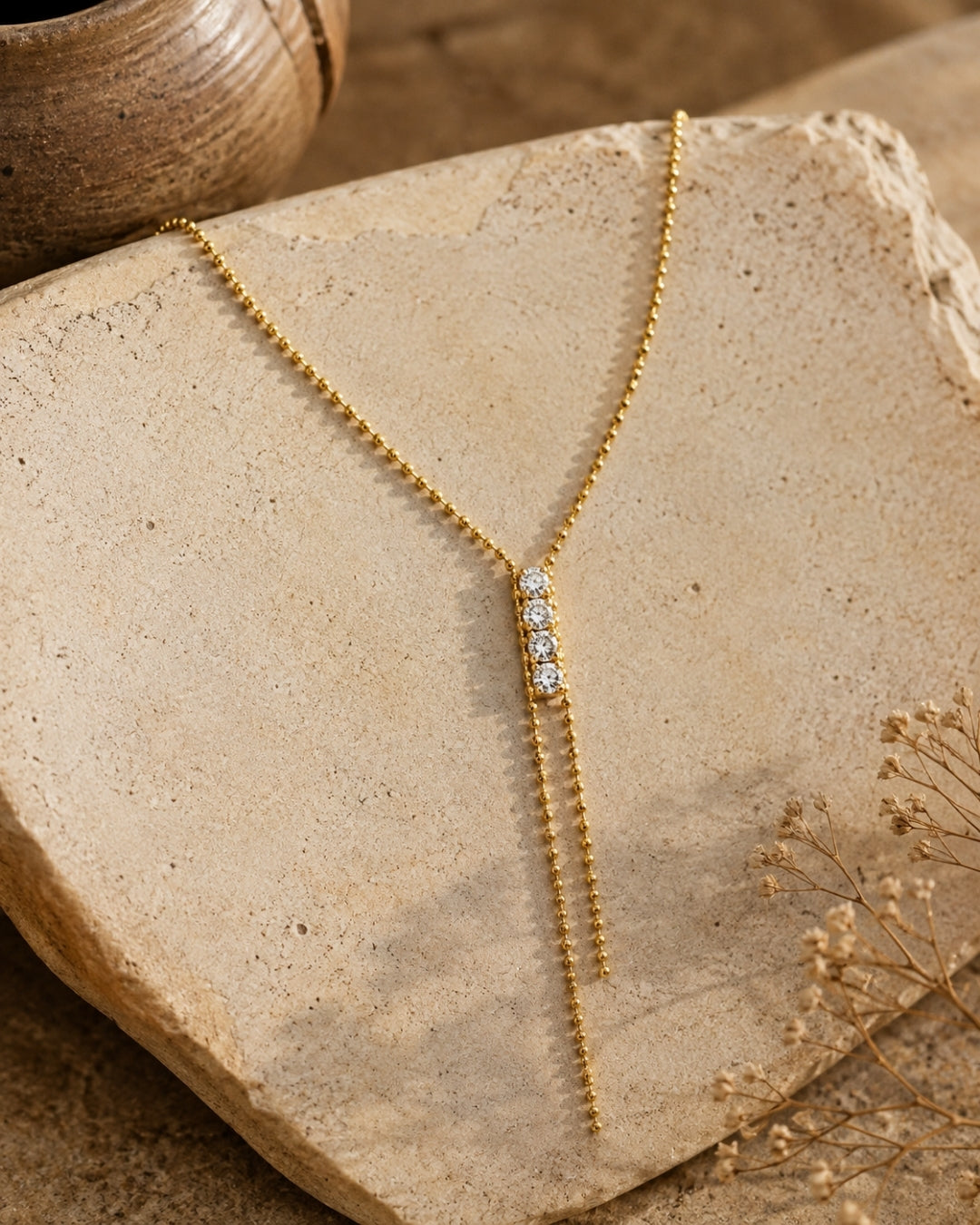 A gold Y-necklace with a central cluster of clear cubic zirconia stones and two dangling chain ends, displayed on a textured beige stone with dried flowers and a ceramic pot in the background.