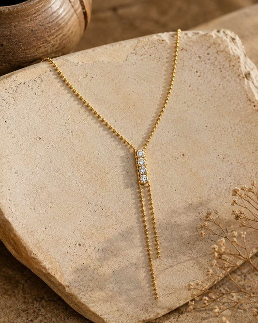 A gold Y-necklace with a central cluster of clear cubic zirconia stones and two dangling chain ends, displayed on a textured beige stone with dried flowers and a ceramic pot in the background.