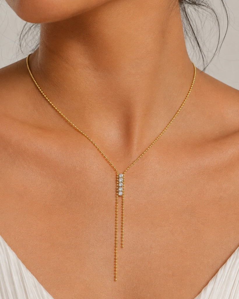 A close-up of a model wearing a delicate gold Y-necklace with a central cluster of small clear stones, paired with a white pleated top against a neutral light gray background.