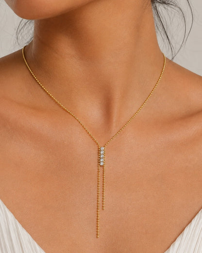 A close-up of a model wearing a delicate gold Y-necklace with a central cluster of small clear stones, paired with a white pleated top against a neutral light gray background.