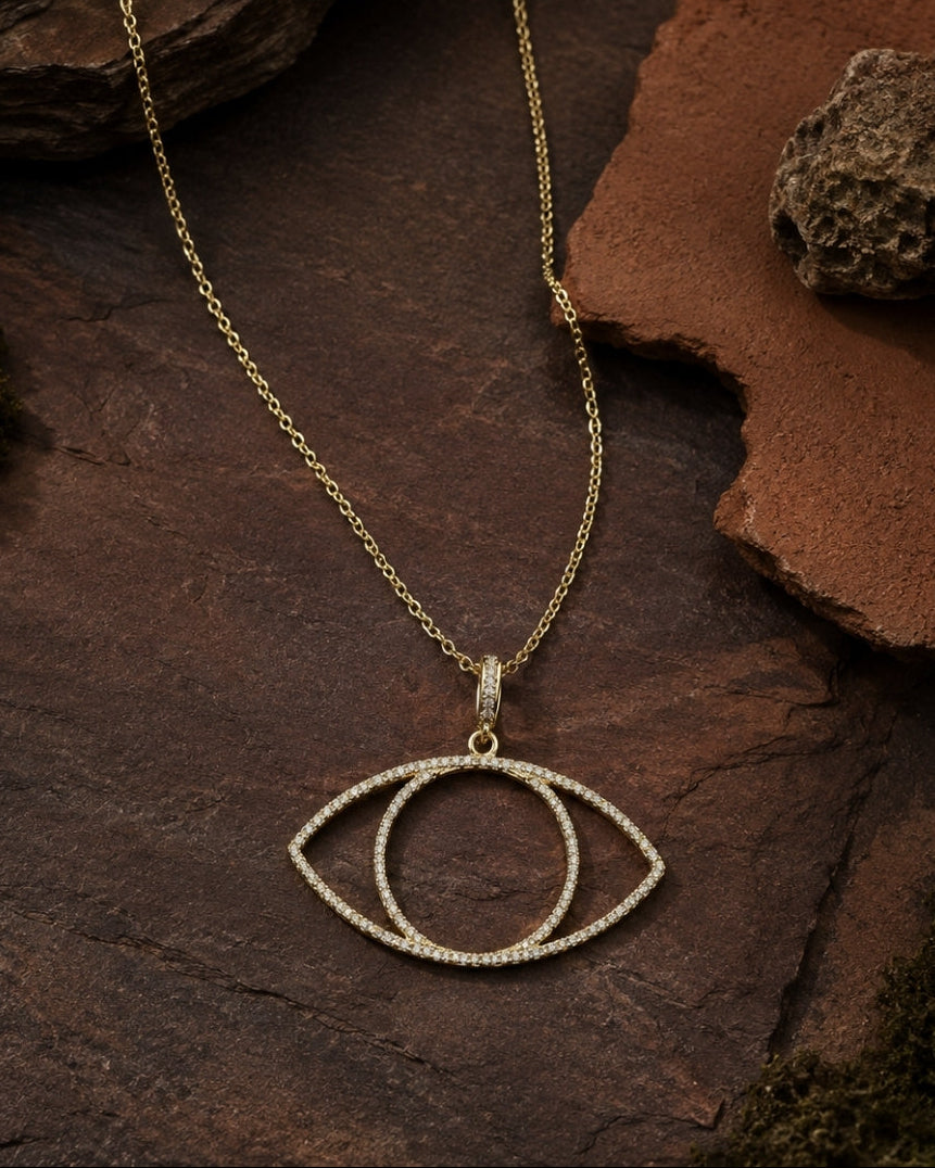 A gold-toned necklace with an eye-shaped pendant set with small clear stones, displayed on a textured dark brown rock surface.