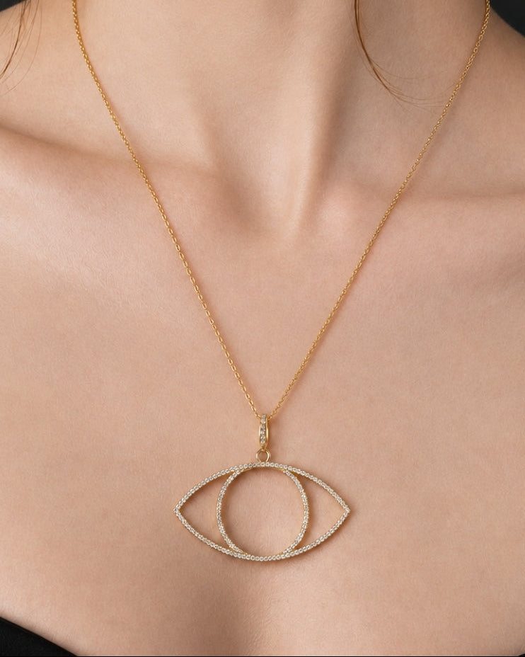 A close-up of a model wearing a delicate gold chain necklace with an eye-shaped pendant studded with small clear crystals, set against a dark background.