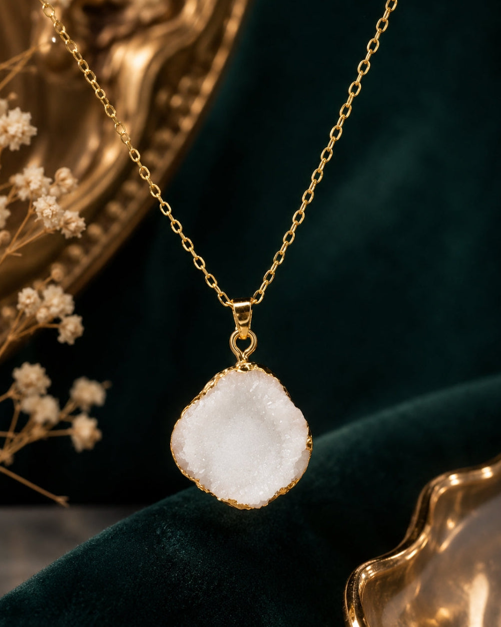A gold chain necklace with a raw, irregular white geode-style pendant, displayed on a dark green velvet surface with gold decorative props.