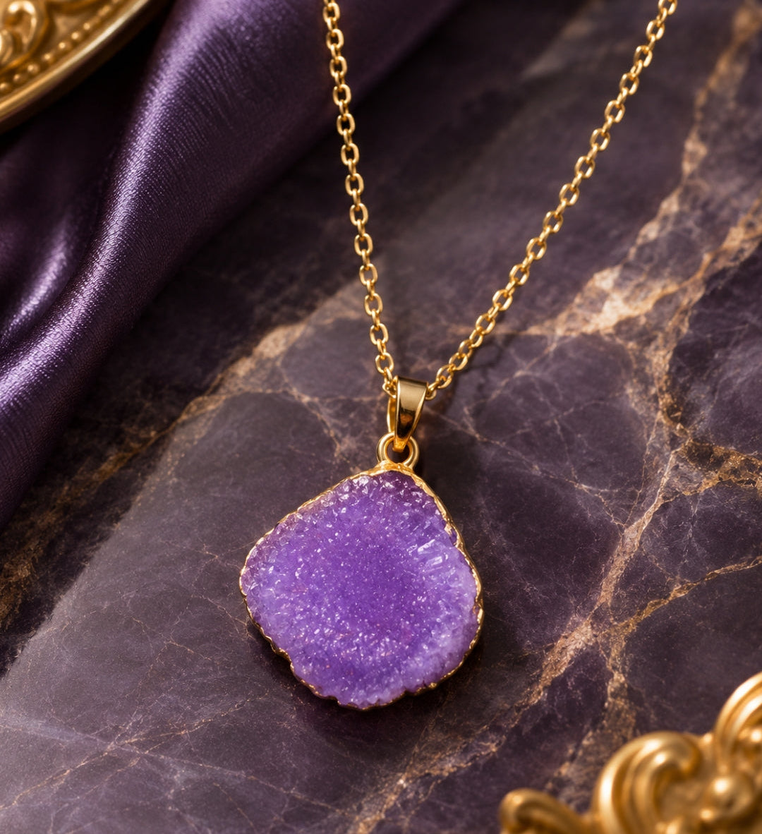A gold chain necklace with an irregular, rough-cut purple geode-style pendant, displayed on a dark purple marble surface with purple satin fabric.