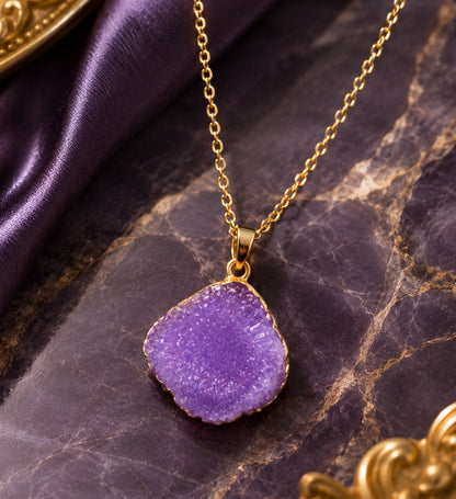 A gold chain necklace with an irregular, rough-cut purple geode-style pendant, displayed on a dark purple marble surface with purple satin fabric.