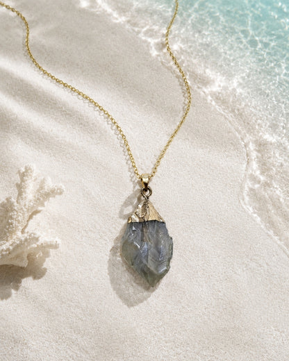 A gold chain necklace with a raw, faceted gray-blue stone pendant, styled on white sand with a piece of coral and ocean water in the background.