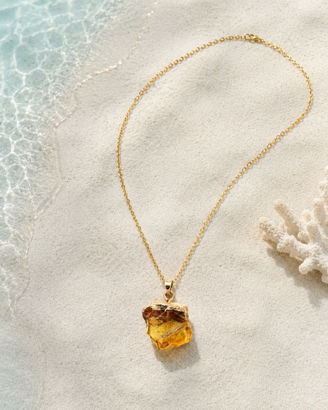 A gold chain necklace with a raw, irregular yellow amber pendant, styled on a white sandy beach with water and a piece of coral in the background.