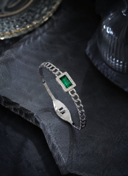A silver chain bangle bracelet featuring a rectangular green emerald-cut center stone surrounded by small clear stones, displayed on a dark textured slate surface.