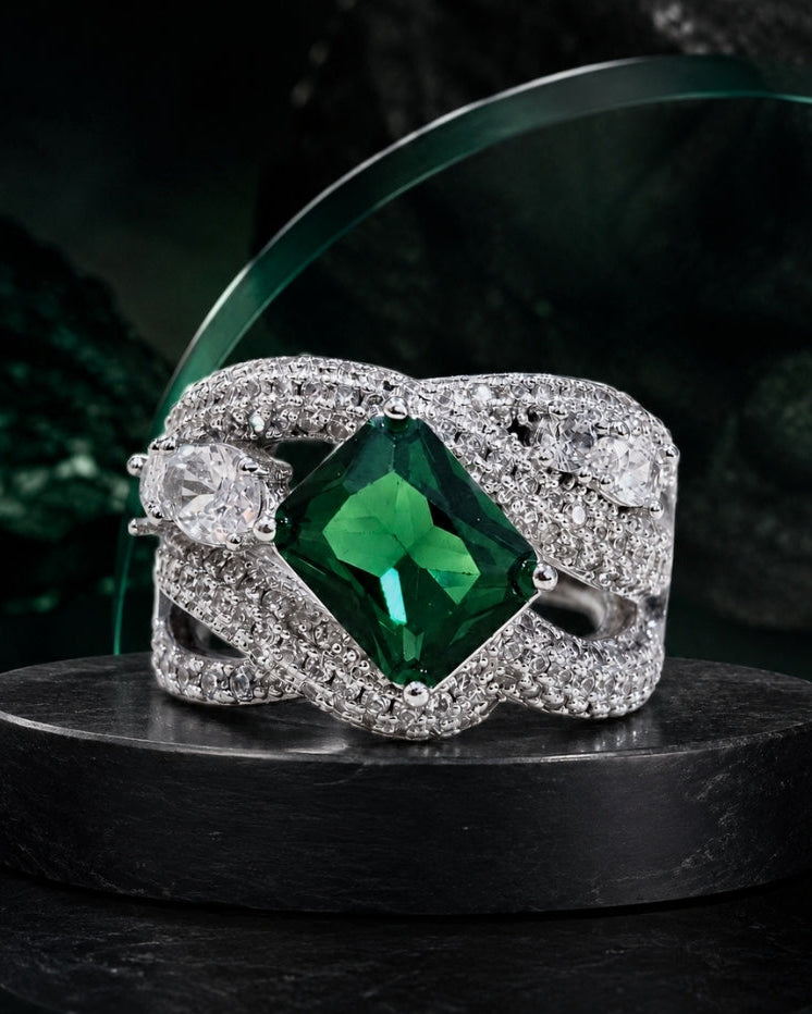 A silver-toned ring with a large square-cut green emerald center stone and pavé-set clear stones in a twisted wrap design, displayed on a dark pedestal.