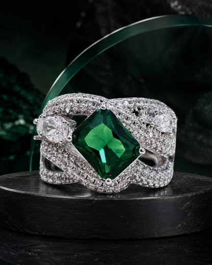 A silver-toned ring with a large square-cut green emerald center stone and pavé-set clear stones in a twisted wrap design, displayed on a dark pedestal.