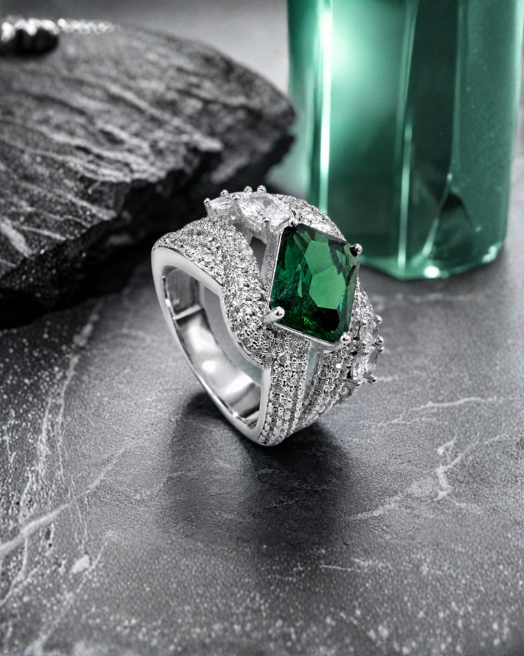 A silver-toned ring featuring a large rectangular green emerald-cut center stone and multiple small clear stones, displayed on a dark textured surface with a green faceted bottle in the background.