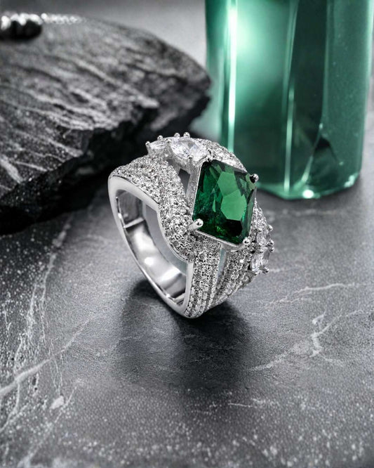 A silver-toned ring featuring a large rectangular green emerald-cut center stone and multiple small clear stones, displayed on a dark textured surface with a green faceted bottle in the background.